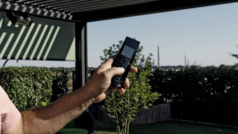 Hand, die eine Fernbedienung hält, mit Blick auf eine Gartenlandschaft und zahlreichen Pflanzen im Hintergrund.
