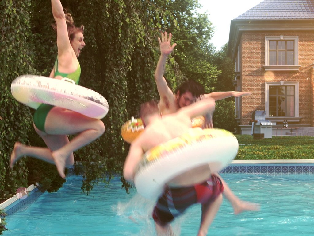 Spritzendes Wasser, vier Personen springen mit Schwimmringen in ein Poolumfeld.