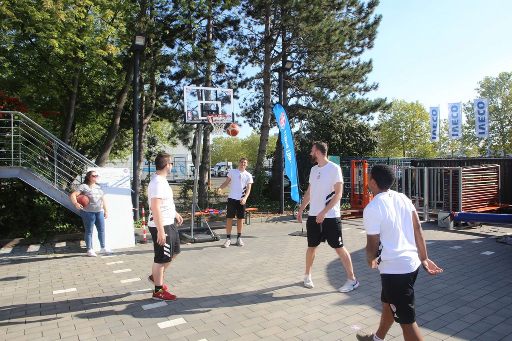 Gruppe von Personen spielt Basketball im Freien, umgeben von Bäumen und einer urbanen Umgebung.