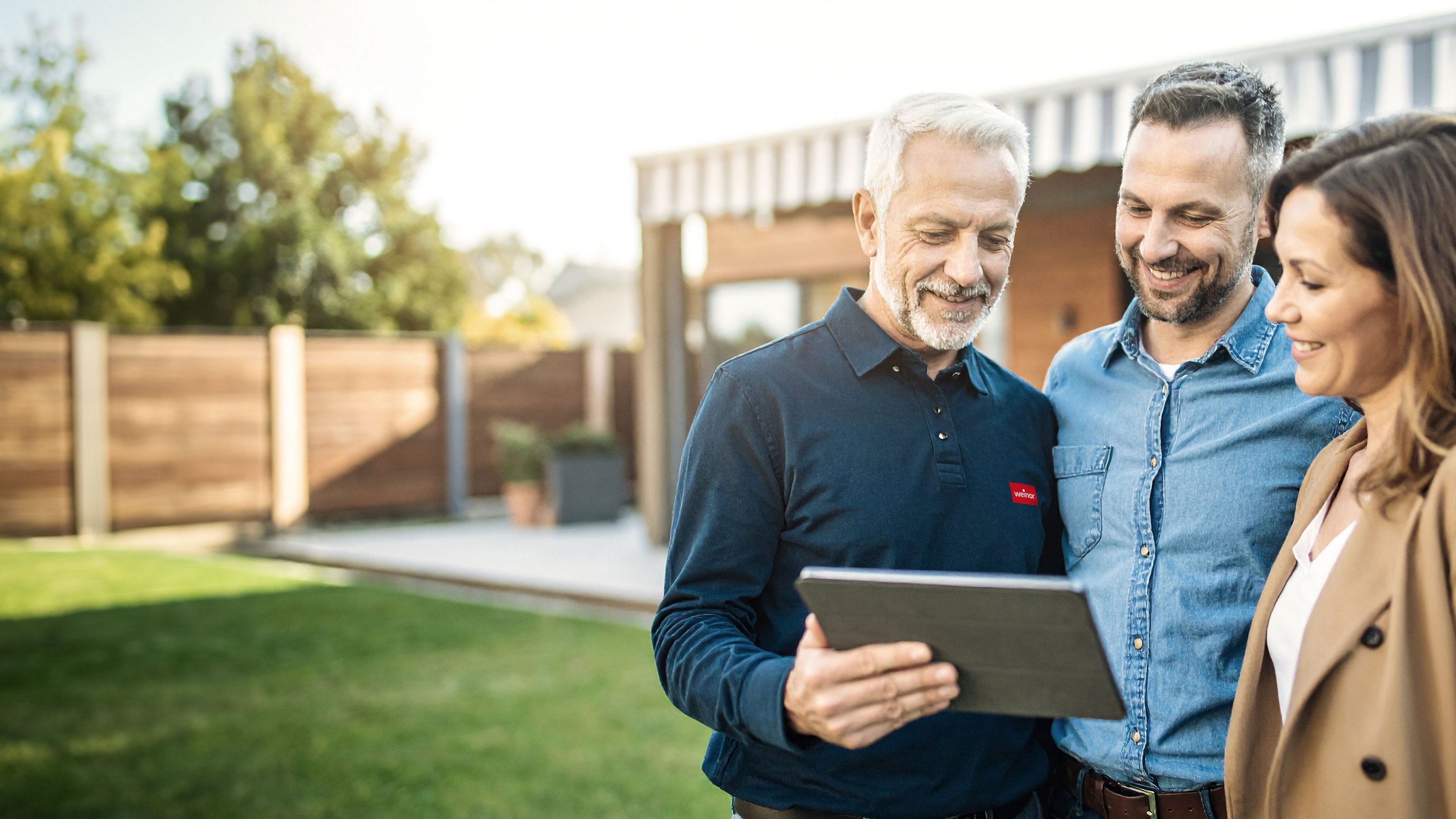 Drei Personen betrachten gemeinsam ein Tablet in einer Terrassensituation mit Überdachung.