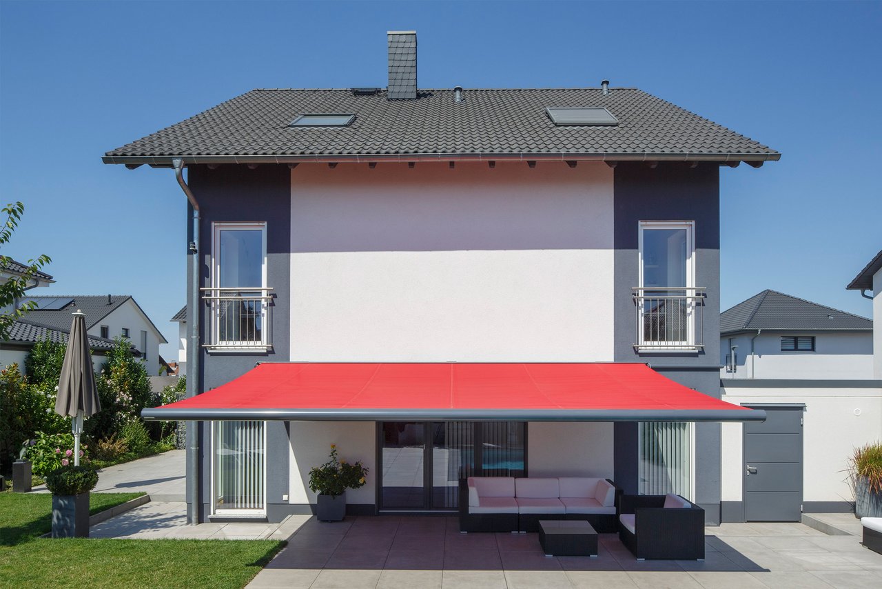 Hausansicht mit roter Markise und Terrassensituation, blauer Himmel im Hintergrund.